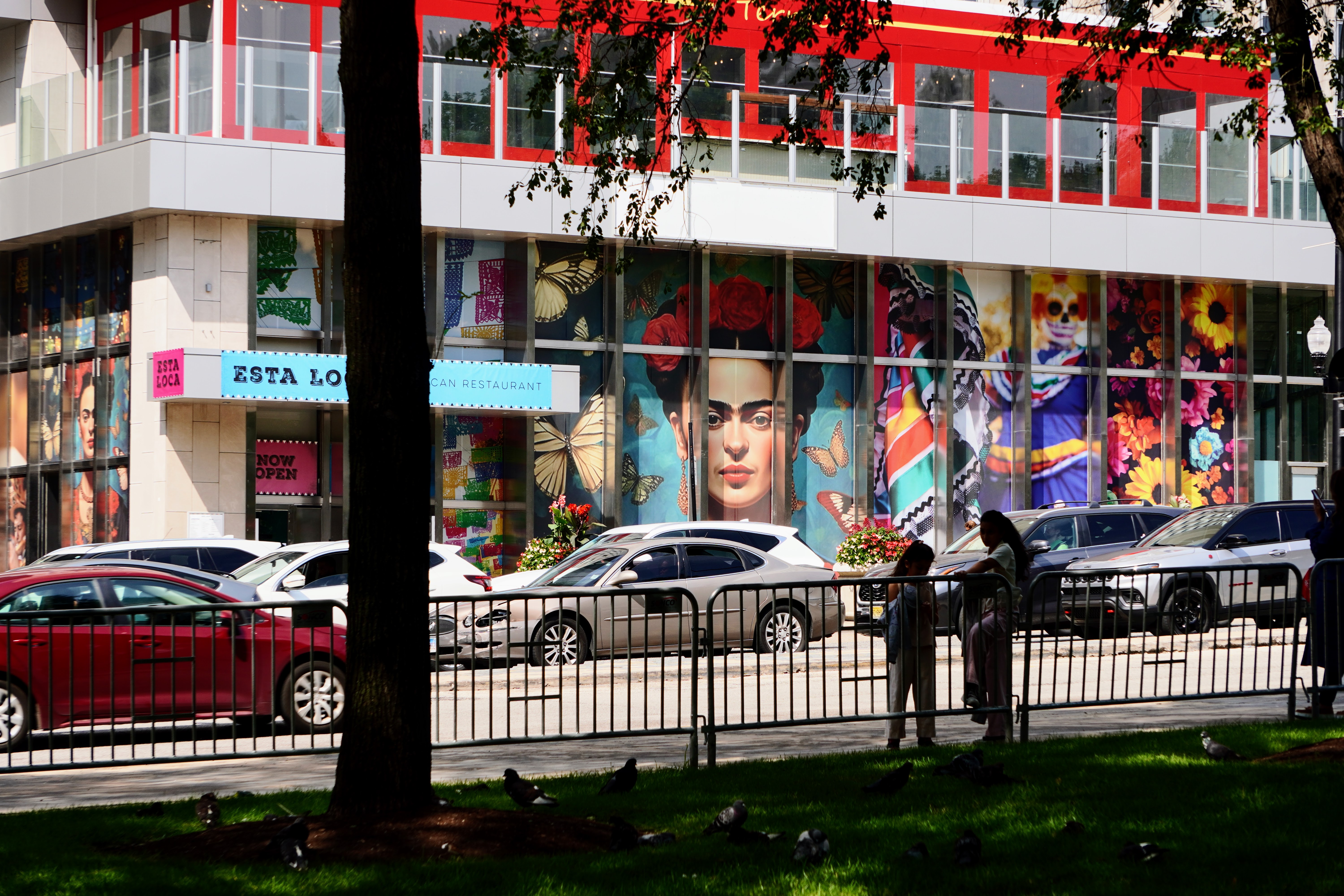 A photo of a street scene in Chicago. There is a Mexican restaurant with a mural of Frida Khalo. People are standing near a fence across the street as cars pass by. The restaurant is part of a larger downtown building.
